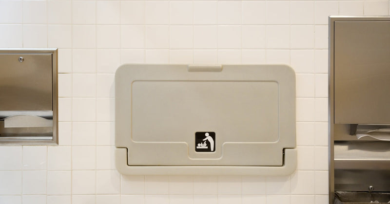 A diaper changing station hangs on the tiled wall of a public bathroom between two paper towel dispensers.