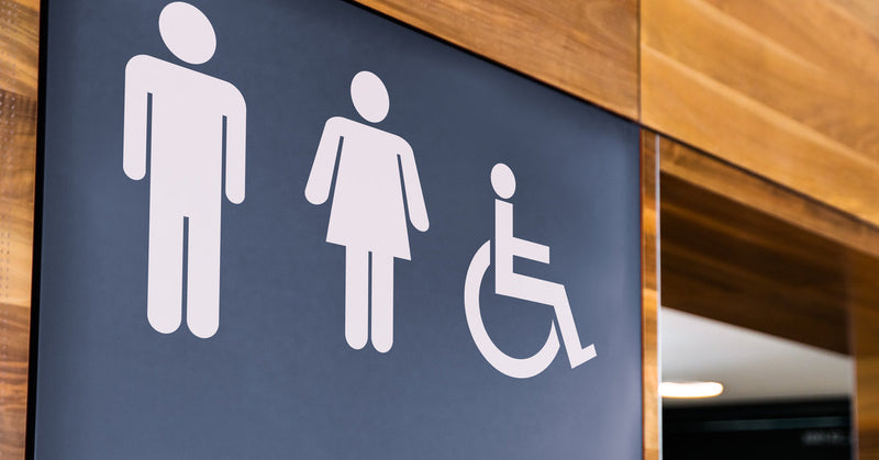 A black public restroom sign with the ADA wheelchair symbol is featured on a wooden wall near a bathroom.