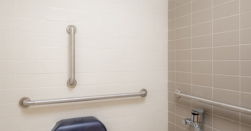 An accessible public restroom features gray tiled walls with three stainless steel grab bars installed near the toilet.s