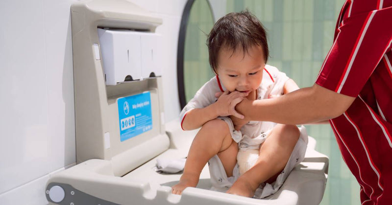 A baby sits up on a commercial diaper changing station, supported by his mother's arms in a public bathroom.