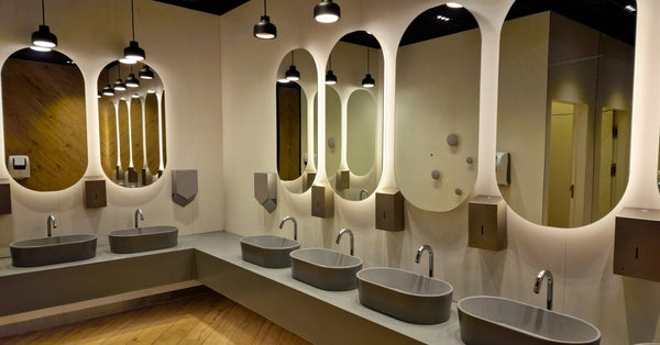 A luxury commercial bathroom with a wood floor features oval mirrors and pendant lighting above gray vessel sinks.