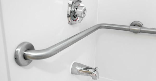 An L-shaped stainless steel grab bar is installed in a clean, white shower stall with a chrome faucet.