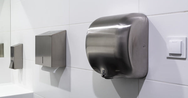 A stainless steel automatic hand dryer hangs on the wall near a paper towel dispenser in a commercial bathroom.