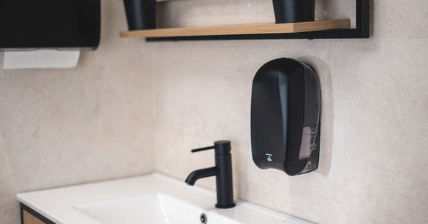 A small black automatic hand soap dispenser is installed on the wall above the sink in a modern bathroom.