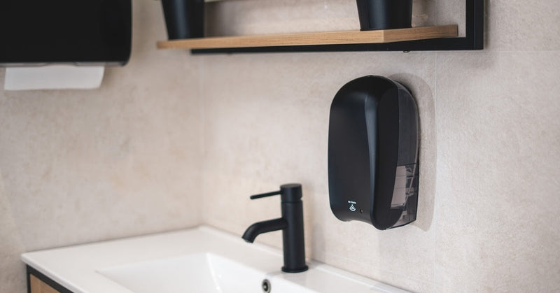 A small black automatic hand soap dispenser is installed on the wall above the sink in a modern bathroom.
