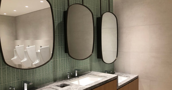 Three oval mirrors sit above three sinks in a modern public restroom. A row of urinals is visible in the mirror's reflection.