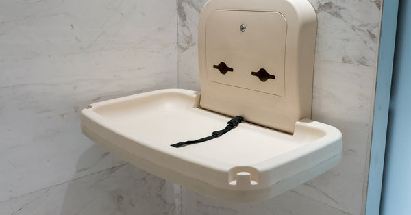 A cream-colored baby changing station hangs open on a white and gray marble wall in a public restroom.
