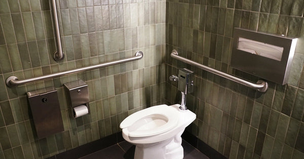A public restroom contains a white toilet, several grab bars, a silver toilet paper holder, and olive green tile on the walls.