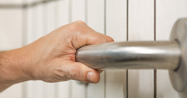 A close-up view shows a hand on a stainless steel grab bar attached to a wall. The wall has white tile.