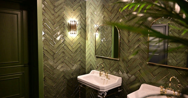 A luxurious restroom features green-tiled walls, two white sinks with gold fixtures, and two mirrors illuminated from the back.