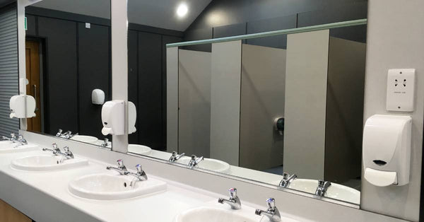 The sink area of a public restroom with two large mirrors on the wall. Three wall-mounted soap dispensers are on the sides.