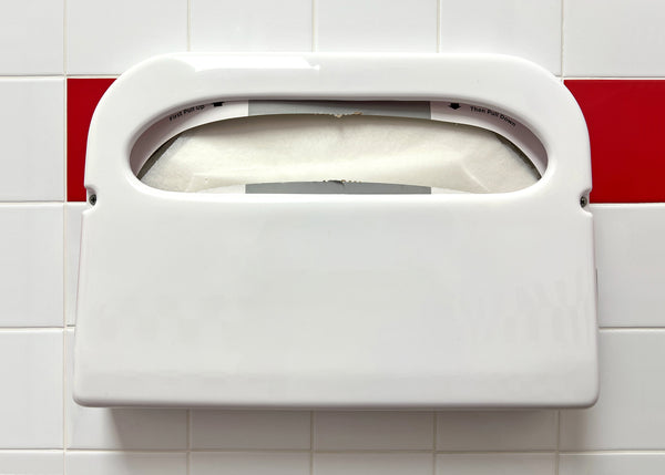 A wall-mounted toilet seat cover dispenser. It's connected to white tiles with a row of red tiles behind it.