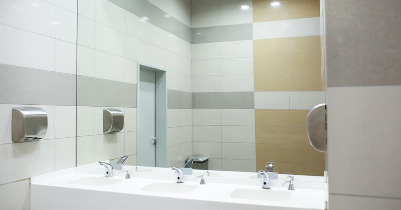 The sink station of a public restroom. A large square mirror takes up the entire wall space behind the sinks.