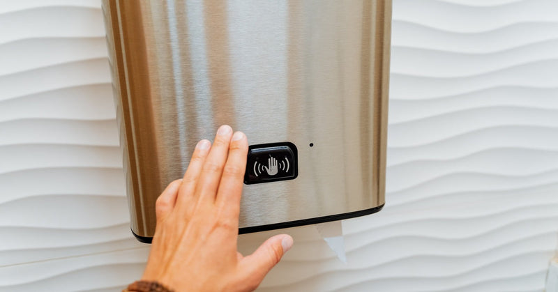 A hand wearing a brown bracelet is reaching out toward a paper towel dispenser with a hand-activated sensor on it.
