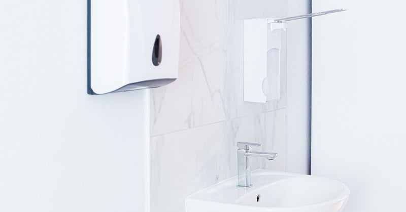 A white wall-mounted automatic soap dispenser on the wall of a public bathroom next to a sink basin.
